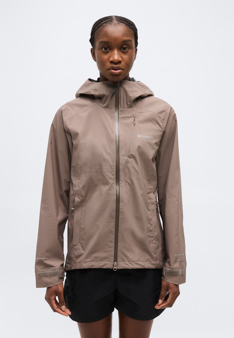 Femme portant une veste de pluie Columbia taupe avec capuche, debout droit devant un fond clair uni.