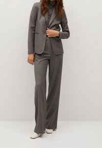 Grijze pak met een slim-fit blazer met notch revers en brede pijpen. Glad materiaal met een subtiel patroon. Gecombineerd met witte puntige schoenen.