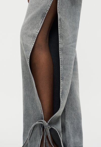 Κοντινό πλάνο ενός ποδιού που καλύπτεται μερικώς από γκρι τζιν παντελόνι με πλευρική σχισμή δεμένη στον αστράγαλο, αποκαλύπτοντας σκούρο δέρμα από κάτω.