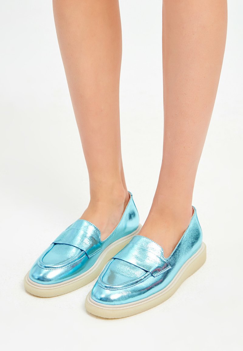 Metallic blaue Slipper mit flacher, weißer Sohle, ausgestattet mit einer glatten Textur und runder Spitze, verziert mit einer gesteppten Naht.