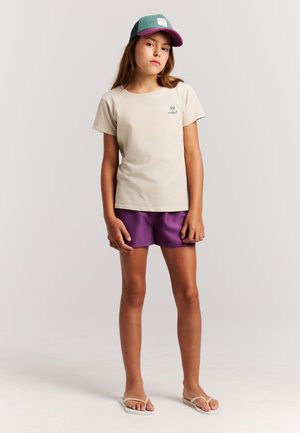 T-shirt en coton beige avec un petit motif de palmier vert, associé à un short violet. Une casquette turquoise et violette ainsi que des tongs blanches complètent la tenue.