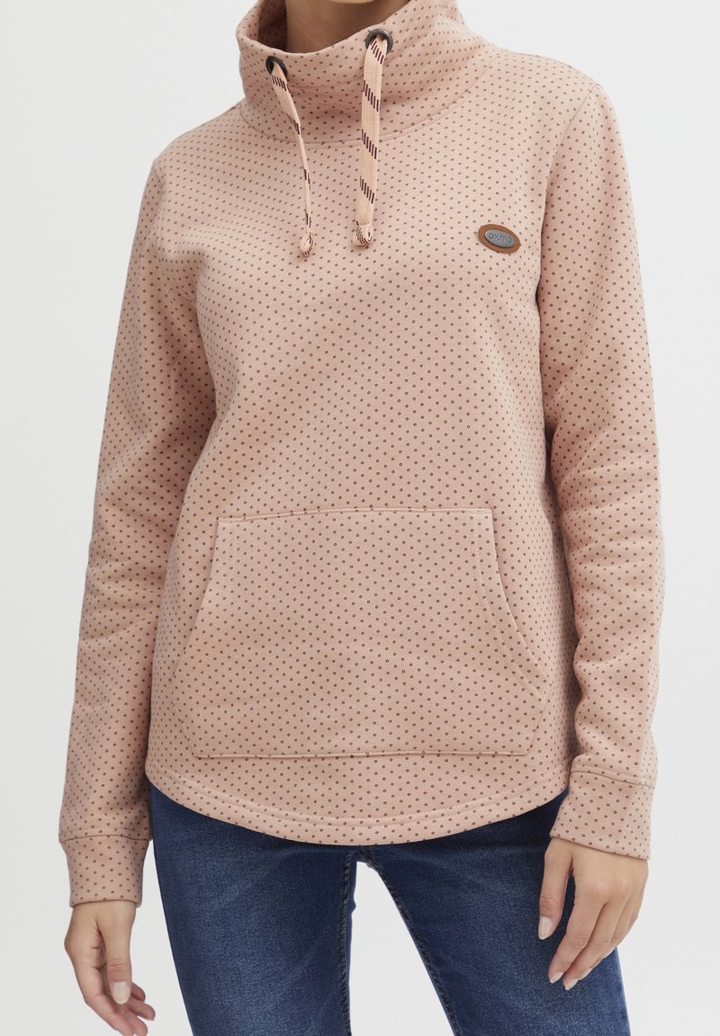 Sweat à capuche rose clair avec motif à pois, col montant, cordons de serrage et poche avant. Texture douce et design à ourlet arrondi.