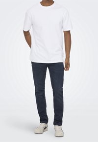 Camiseta blanca de algodón con cuello redondo clásica, combinada con jeans de mezclilla azul oscuro, complementada con zapatillas de color claro.