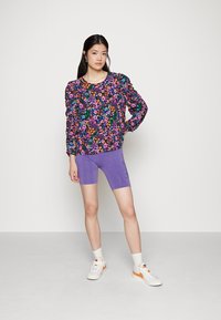 Blusa de manga longa estampada com flores e mangas bufantes em púrpura, combinada com calções púrpura e ténis brancos com detalhes em laranja.