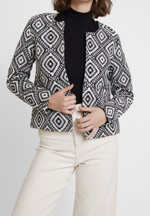 Femme portant une veste à motif géométrique noir et blanc sur un col roulé noir et un pantalon crème, ajustant la veste avec les deux mains.