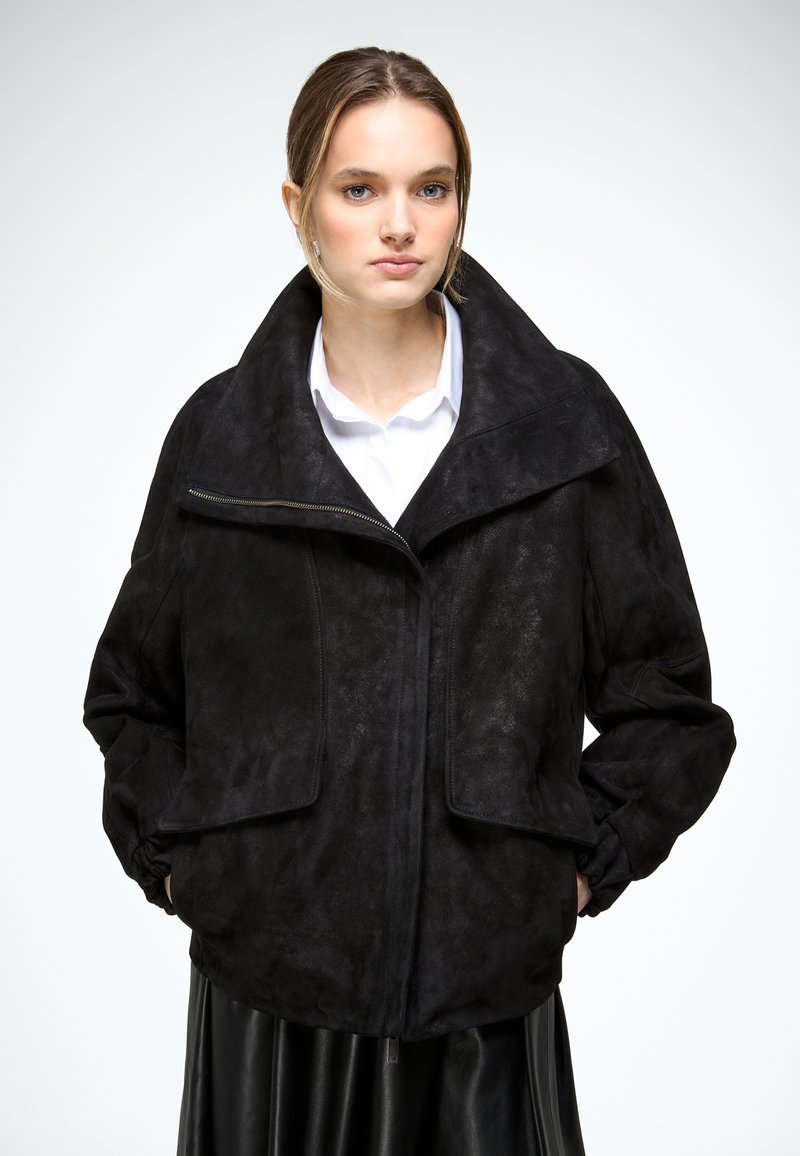 Jeune femme portant une grande veste noire surdimensionnée avec un large col et une fermeture éclair frontale, debout les mains dans les poches sur un fond blanc.