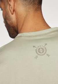 Světle zelené tričko s kulatým výstřihem, které má na zadní straně design loga se směrovými šipkami a textem "NORTH".