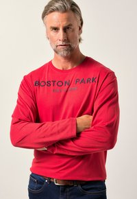 Rotes Langarm-T-Shirt mit dem Aufdruck "BOSTON PARK EST 1996" in kontrastierendem Dunkelblau, kombiniert mit blauen Jeans und einem braunen Gürtel.