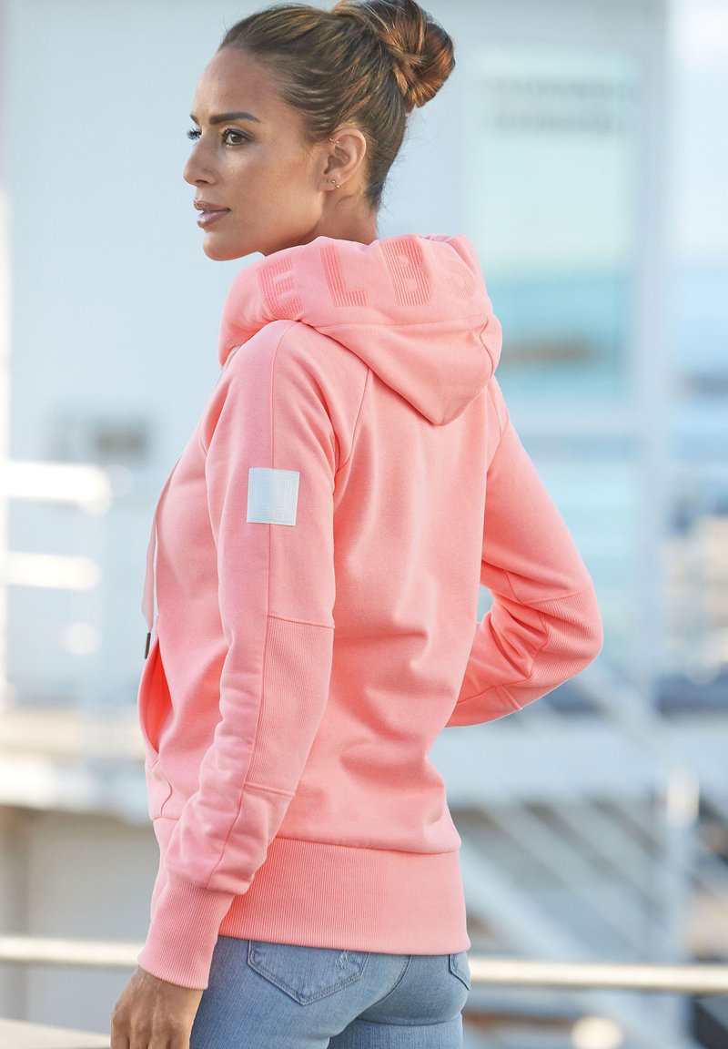 Pink hoodie with a large hood, ribbed cuffs, and a white patch on the left arm. Features subtle lettering along the hood edge. Soft texture.