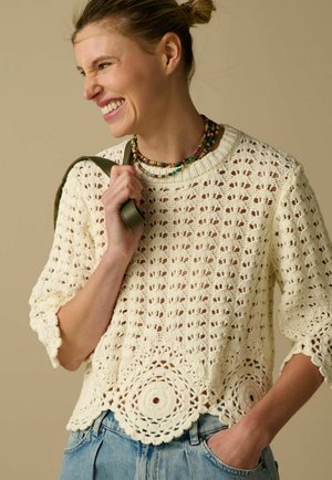 Femme souriante portant un pull en crochet crème avec un ourlet festonné et des colliers multicolores en perles, tenant une sangle verte sur son épaule.