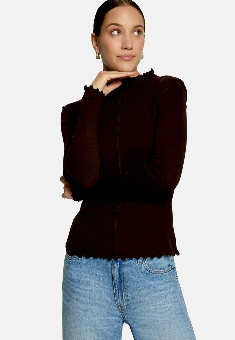 Femme portant un haut à manches longues marron foncé et un jean bleu clair, main reposant sous le menton, regardant sur le côté, portant des boucles d’oreilles créoles dorées.