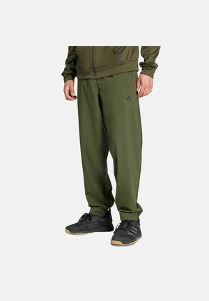 Mann trägt olivgrüne Adidas-Trainingshose und passende Jacke, dazu schwarze Sportschuhe vor hellem Hintergrund.