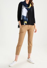 Schwarzes Langarmoberteil, beiges tailliertes Hose und ein mehrfarbiges Schal mit navy, gelben und weißen Abschnitten. Weiße Sneaker vervollständigen das Outfit.