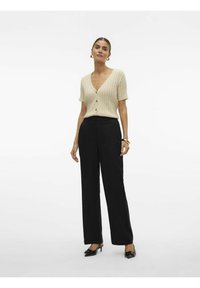 Cardigan beige à manches courtes et côtelé avec des boutons, associé à un pantalon noir à jambes larges et des talons aiguilles noirs, sur fond blanc.
