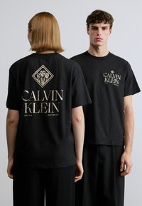Μαύρα βαμβακερά μπλουζάκια με χρυσό λογότυπο «CALVIN KLEIN» εμπρός και πίσω, με γραφικό διαμάντι λουλουδιών στο πίσω μέρος. Κοντά μανίκια, άνετη εφαρμογή.