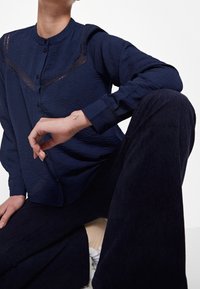 Camicia blu marina con superficie testurizzata, caratterizzata da inserti in pizzo sul petto e un colletto classico; abbinata a pantaloni in velluto a coste scuri.