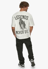 Fehér, oversized póló fekete "LEGENDS NEVER DIE" felirattal és imádkozó kezek grafikájával a hátsó részen. Fekete cargo nadrág és sportcipő.