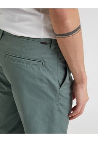 Muž v zelených kalhotách a bílém tričku, ukazuje tetovaný předloktí se dvěma černými pásky blízko lokte.