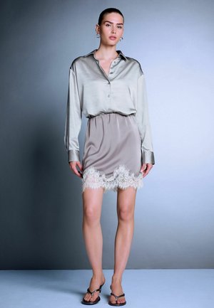 Woman standing wearing a light gray satin shirt tucked into a taupe satin skirt with white lace trim and black heeled sandals on a plain background.