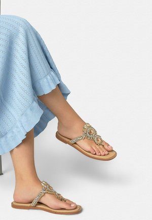 Piernas cruzadas con pantalones de punto azul claro y sandalias planas beige decoradas con adornos florales dorados y de pedrería.