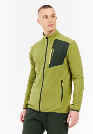 Man in een groene ritsjack met een zwarte borstzak en bijpassende broek, kijkt naar rechts tegen een witte achtergrond.