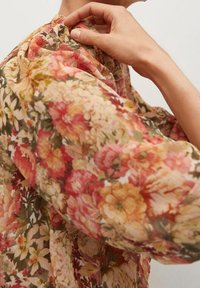 Een hand die een knoop vastmaakt op een blouse met hoge hals en lange mouwen, met een dicht bloemmotief in warme tinten.