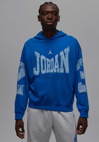 Niebieska bluza z kapturem z napisem "JORDAN" w białym kolorze, kieszenią kangurka oraz małym białym logo na piersi; noszona z szarymi spodniami dresowymi.
