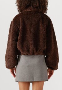 Bruine, fluffige jas met hoge kraag en elastische tailleband, gecombineerd met een korte grijze rok, gezien vanuit de achterkant.