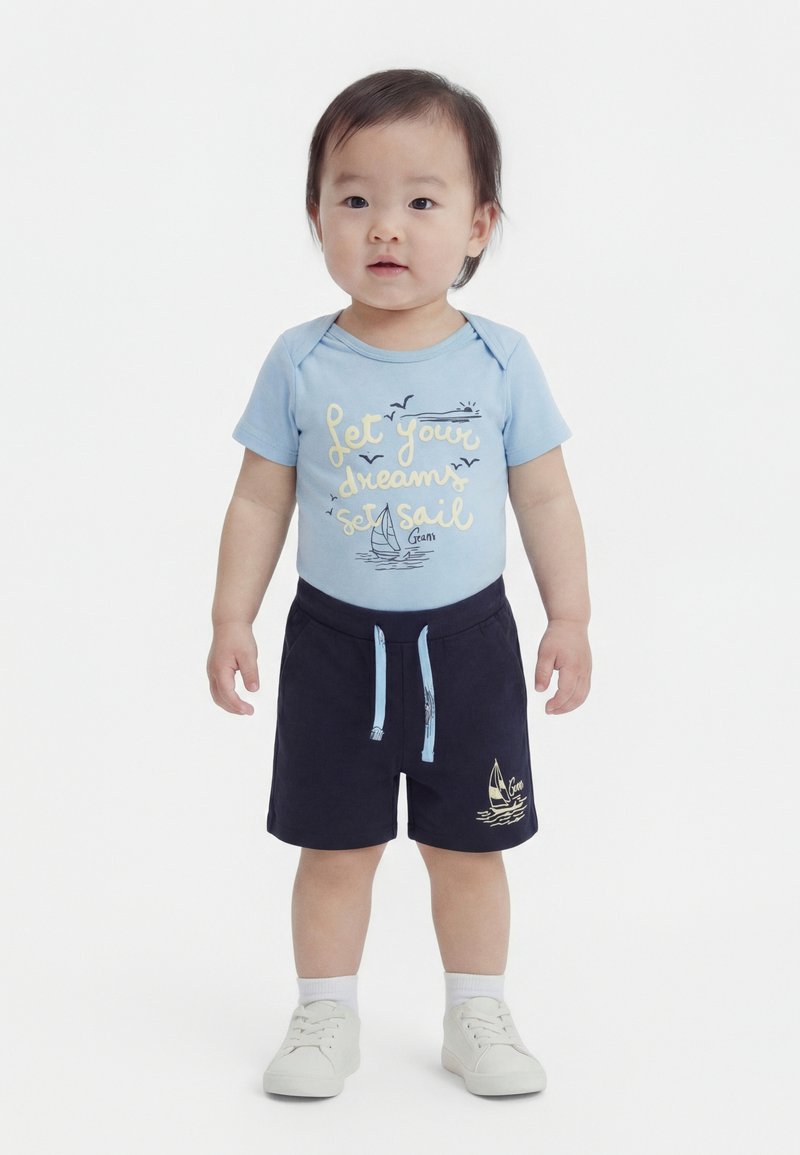 Νήπιο με ανοιχτό μπλε πουκάμισο με το κείμενο «Άσε τα όνειρά σου να σαλπάρουν» και σκούρο μπλε σορτς με σχέδιο ιστιοφόρου, στέκεται μπροστά σε λευκό φόντο.