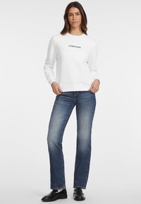 Valkoinen huppari, jossa on musta "GUESS JEANS" -logo, yhdistettynä sinisiin farkkuihin ja mustiin loafereihin. Neutraali tausta, rento tyyli.