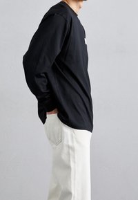 Sort langærmet skjorte med rund hals, vist sammen med hvide denimbukser med sidesømme og detaljerede syninger på baglommen.