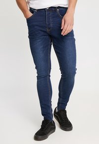 Mörkblå skinny jeans med ljusa sömmar, femfickorsdesign och knappgylf. Bärs med svarta sneakers mot en neutral bakgrund.