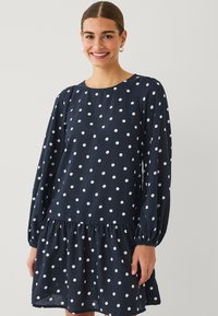 Vestido de lunares en color navy con mangas largas y dobladillo fruncido, hecho de tela ligera. Presenta un escote redondo y patrones circulares blancos.
