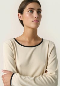 Maglietta beige a manica lunga con texture a coste, bordi neri attorno al collo e alle maniche, e un fit rilassato.