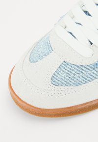 Sandači od antilopa i trapera s bijelim gornjim dijelom, svijetloplavim denim detaljima, ravnim bijelim vezicama i smešim gumiranim đonom. Zakrivljeni dizajnerski elementi.