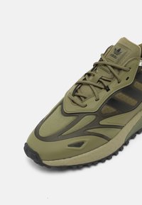 Baskets de sport vert olive en matériaux mesh et synthétiques, dotées d'une semelle texturée, d'un système de laçage et d'accents noirs contrastants.