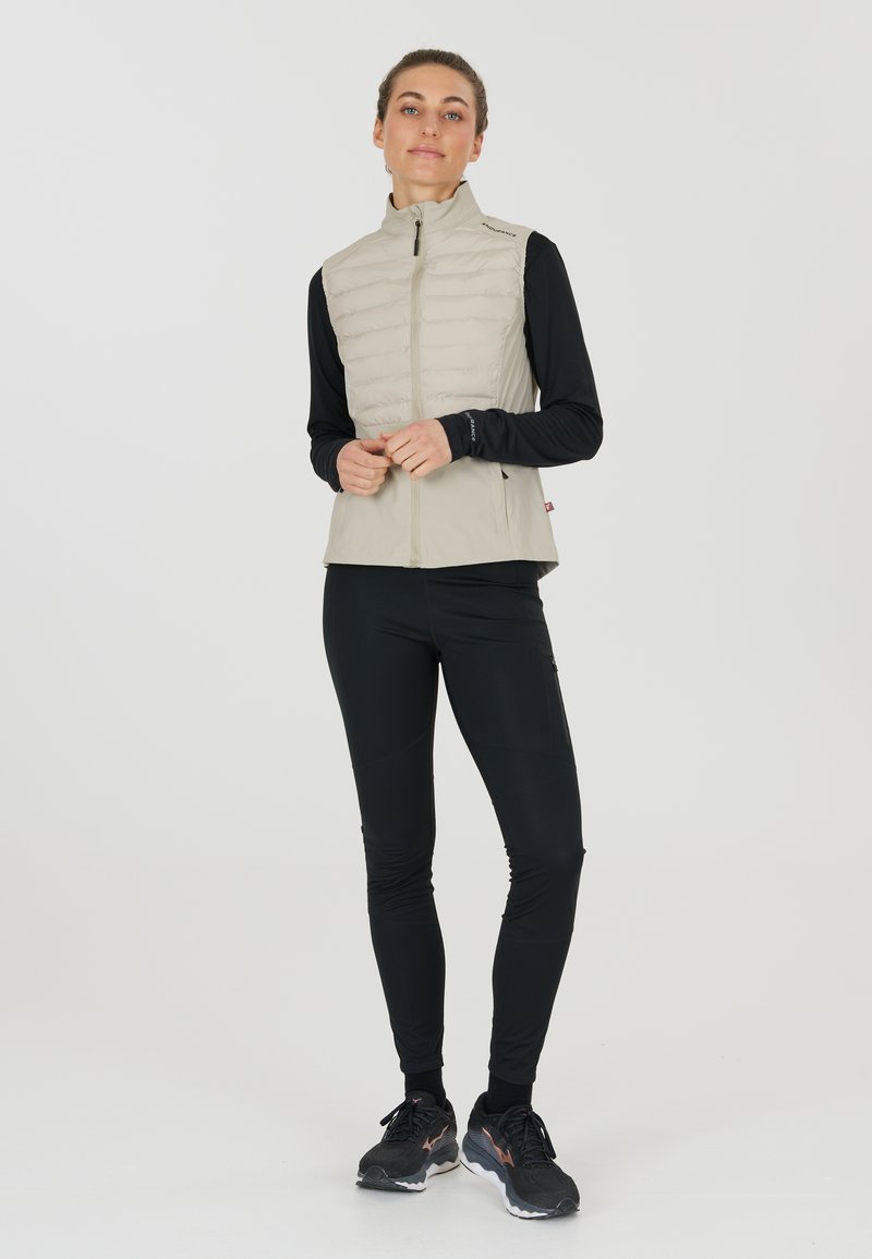 Leichter beiger ärmelloser Weste mit gestepptem Design, kombiniert mit schwarzen langen Ärmeln und enganliegenden Hosen. Verfügt über einen Reißverschluss und sportliche Schuhe.