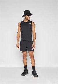 Svart ermeløs topp, matchende shorts og svart bøttehatt. Svarte joggesko med teksturerte såler. Logo vist på skjorten og shortsen.