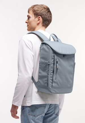 Junger Mann trägt einen hellgrauen rechteckigen Rucksack mit vorderer Reißverschlusstasche und seitlichen Fächern, gekleidet in weißem Hemd und blauer Jeans.