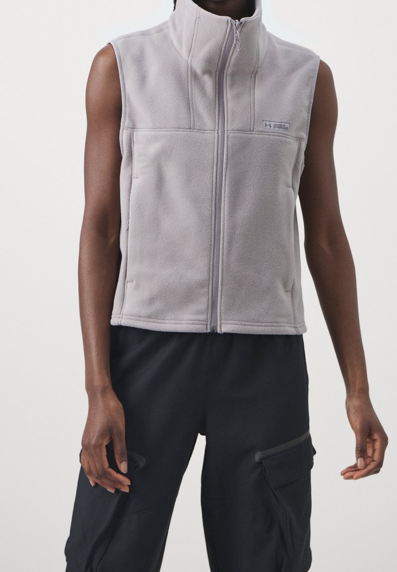 Veste sans manches en polaire gris clair avec fermeture éclair à l’avant portée sur un pantalon cargo noir sur une personne à la peau foncée avec les bras détendus.