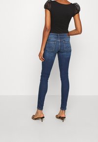 Tunna blå denimjeans med hög midja, diskreta fickor och en texturerad baksidestyk. Matcherade med öppna högklackade skor.