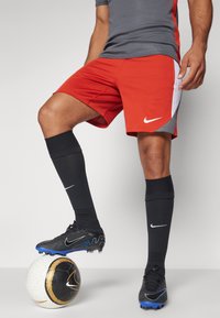 Pantalones cortos deportivos rojos con acentos en blanco y gris, calcetines negros y botas negras, de pie sobre un balón de fútbol blanco con detalles dorados.
