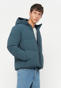 Teal puffer jacket with a hood, zip closure, and side pockets. Features quilted texture, lightweight fabric, and a relaxed fit.