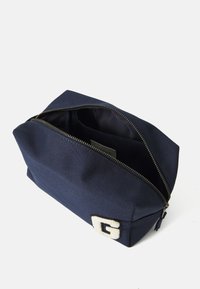 Pochette en toile bleu marine avec une finition texturée, ornée d'un logo "G" brodé en blanc et d'une fermeture éclair dorée. L'intérieur comprend une petite poche.