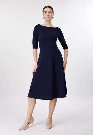Kvinde iført en mørkeblå, knælang kjole med trekvartlange ærmer og beige høje hæle, stående mod en ensfarvet hvid baggrund.
