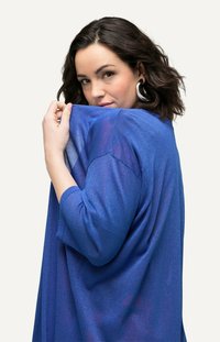 Modrá svetr s volným střihem, tříčtvrtečními rukávy a třpytivou texturou. Modelka je částečně otočená a drží část svetru.