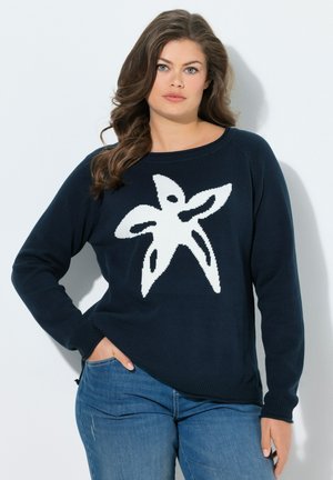 Kvinde med langt brunt hår iført en navyblå sweater med et stort hvidt søstjernemotiv og blå jeans, stående mod en hvid baggrund.