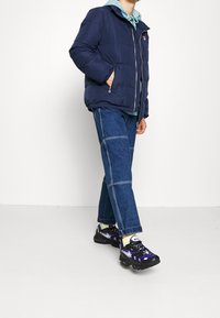 Marinblå puffjacka med dragkedja, bärs över en ljusblå huvtröja. Blå denimbyxor med lappar, kombineras med svarta och lila sneakers.