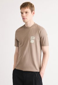 Brun bomulls t-skjorte med korte ermer, rund hals og en liten grafisk logo trykk på venstre bryst. Myk tekstur og avslappet passform.
