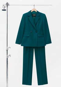 Szmaragdowy, podwójnie zapinany żakiet z wyłogami i bocznymi kieszeniami, w połączeniu z dopasowanymi spodniami o prostym kroju, wiszący na wieszaku.